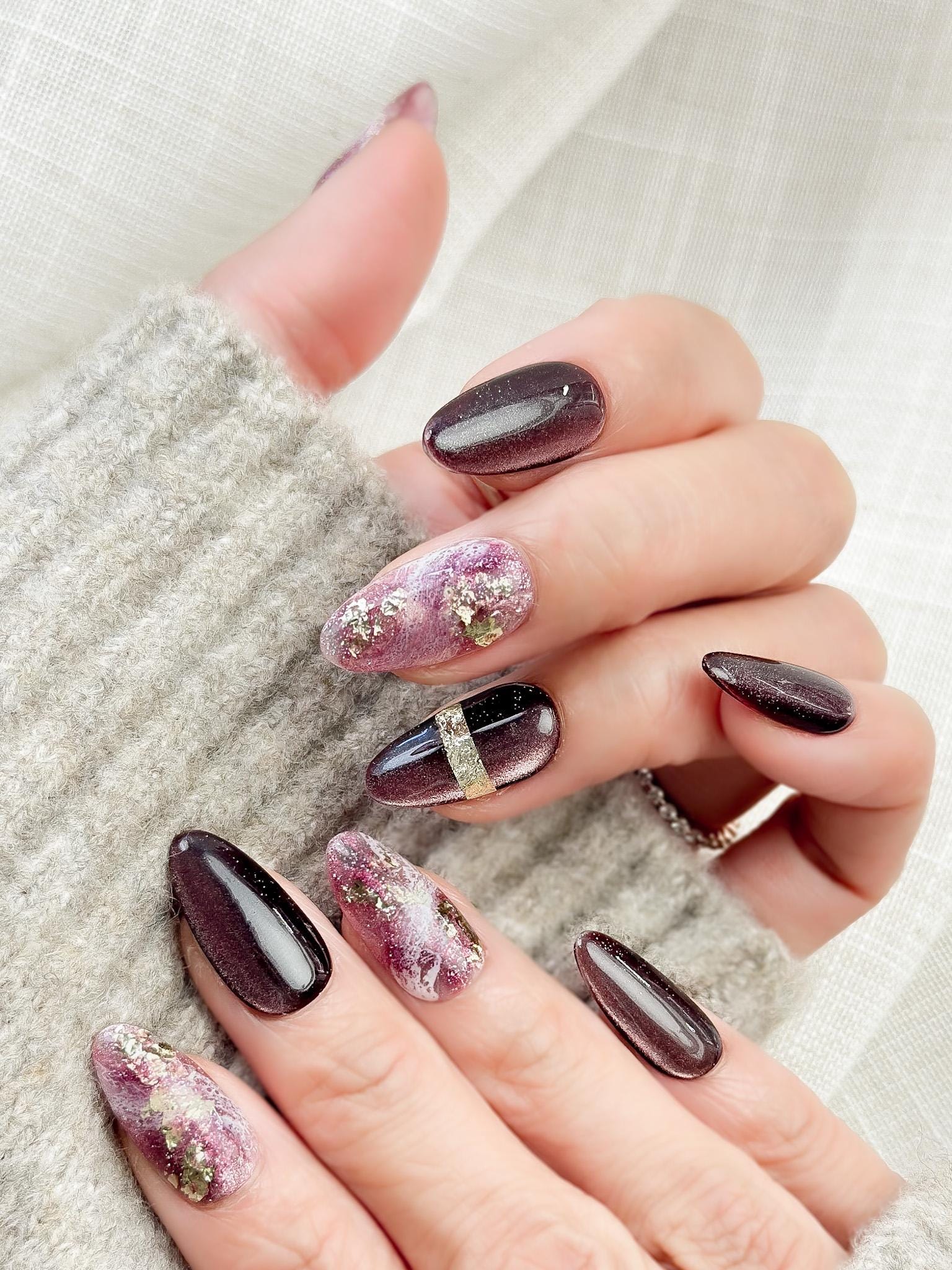 Hand with Almond-shaped press-on nails in deep plum and black cat-eye shimmer with pink marble accents, gold foil detailing, and glossy finish