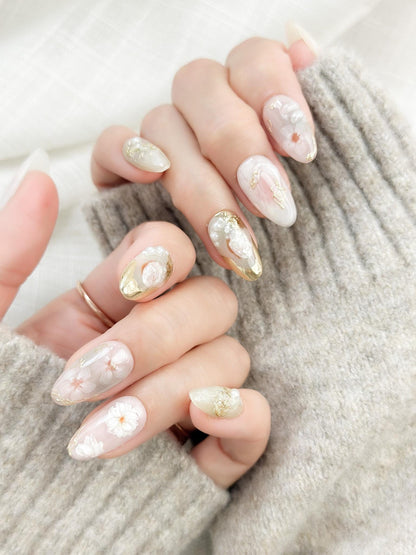 Hand with Almond-shaped white and pearlescent press-on nails with 3D floral accents, gold chrome details, pearl embellishments, and soft milky marble art