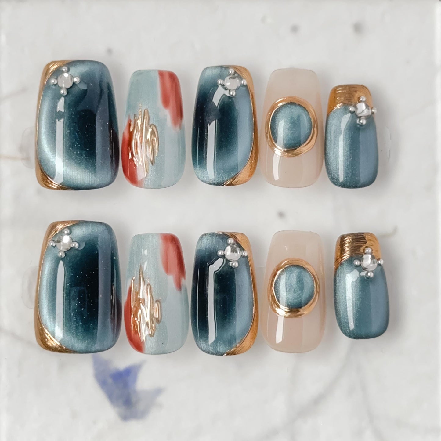 Close-up of deep blue and silver chrome press-on nails with gold accents and gemstone details, featuring a metallic cat-eye shimmer