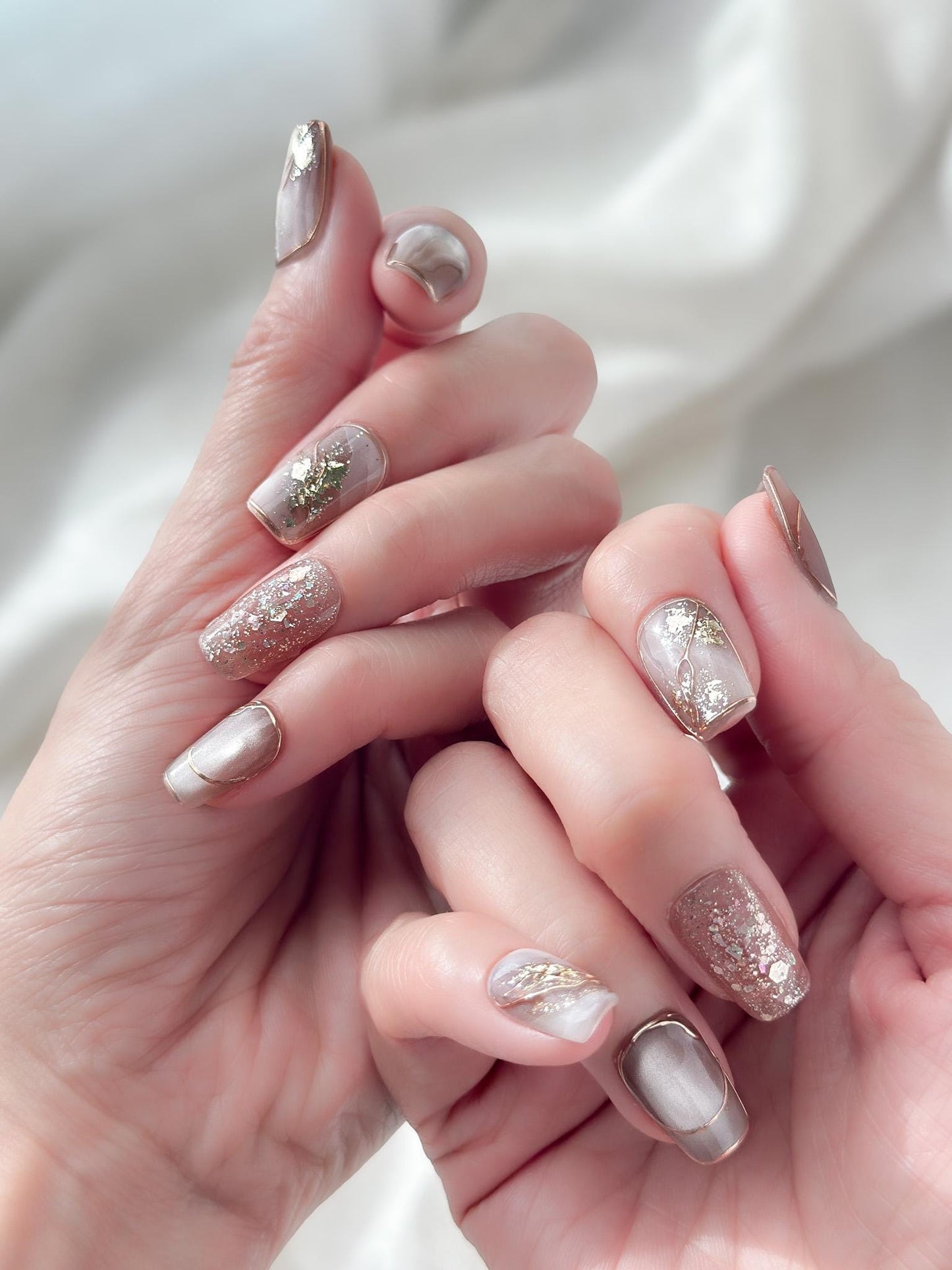Close-up of hands with nude and taupe press-on nails with gold foil accents, rose gold glitter, and marble details