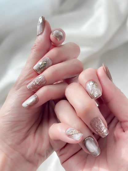 Close-up of hands with nude and taupe press-on nails with gold foil accents, rose gold glitter, and marble details