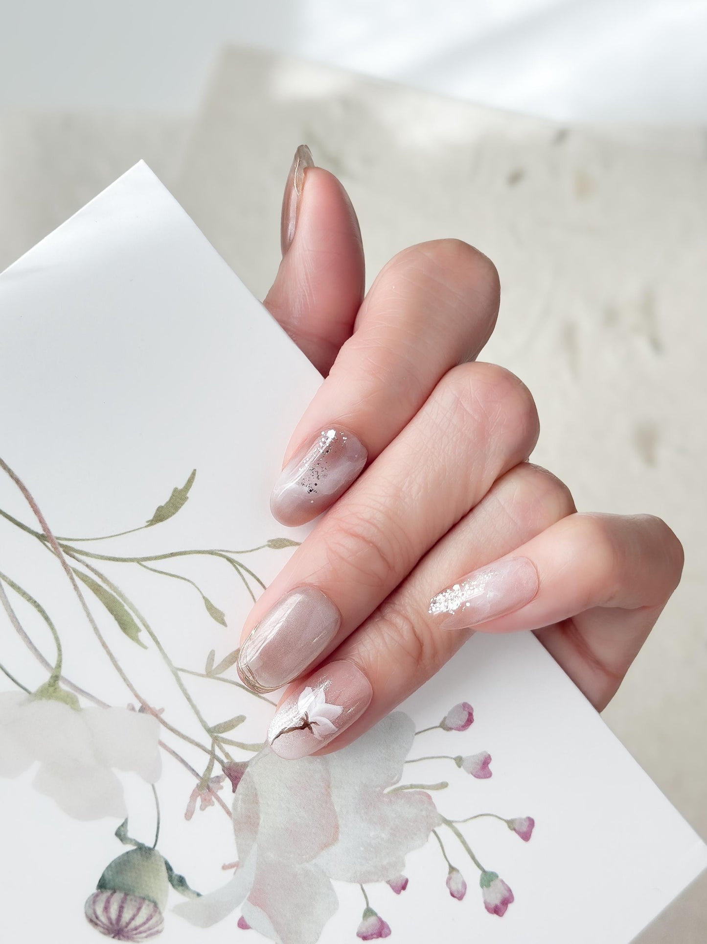 Hand with Pink and pearl white press-on nails with soft marble accents, hand-painted white floral art, silver glitter details, and glossy finish