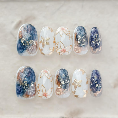 Oval-shaped press-on nails with deep navy and gold marble art, gold foil accents, and soft floral details.