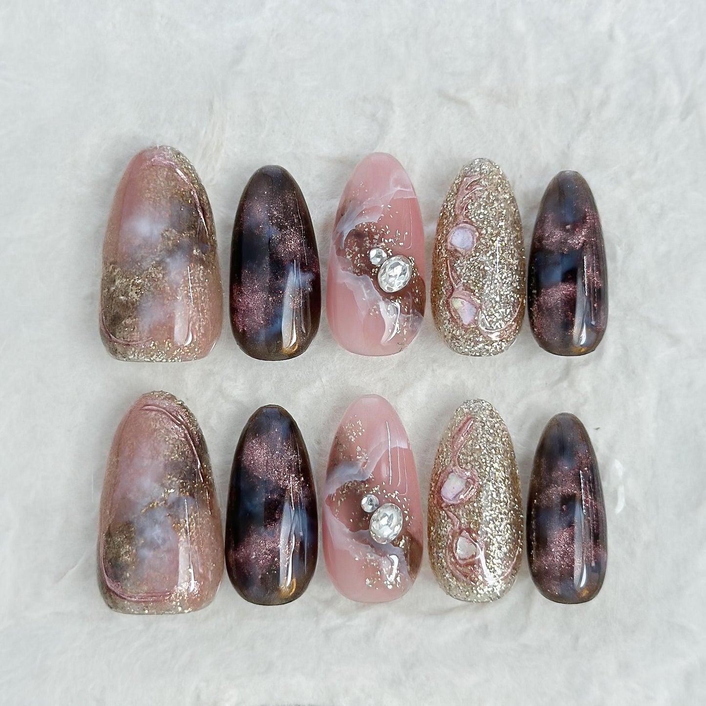 Almond-shaped luxury press-on nails in metallic brown and black cat-eye shimmer with pink marble accents, gold glitter, and pearl gemstone details