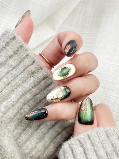 Hand with Green and gold gemstone-inspired press-on nails with cat-eye shimmer, glitter accents, gold foil detailing, and 3D crystal embellishments