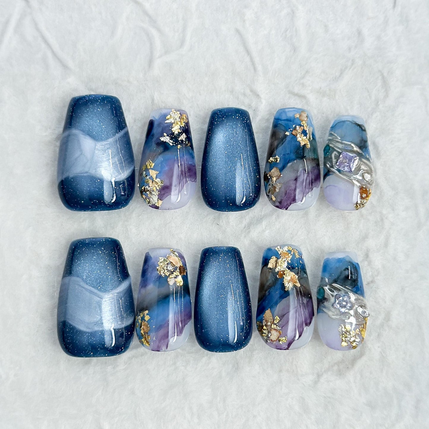 Blue galaxy press-on nails with starry cat-eye shimmer and watercolor marble accents in navy, lavender, and white, finished with gold foil details
