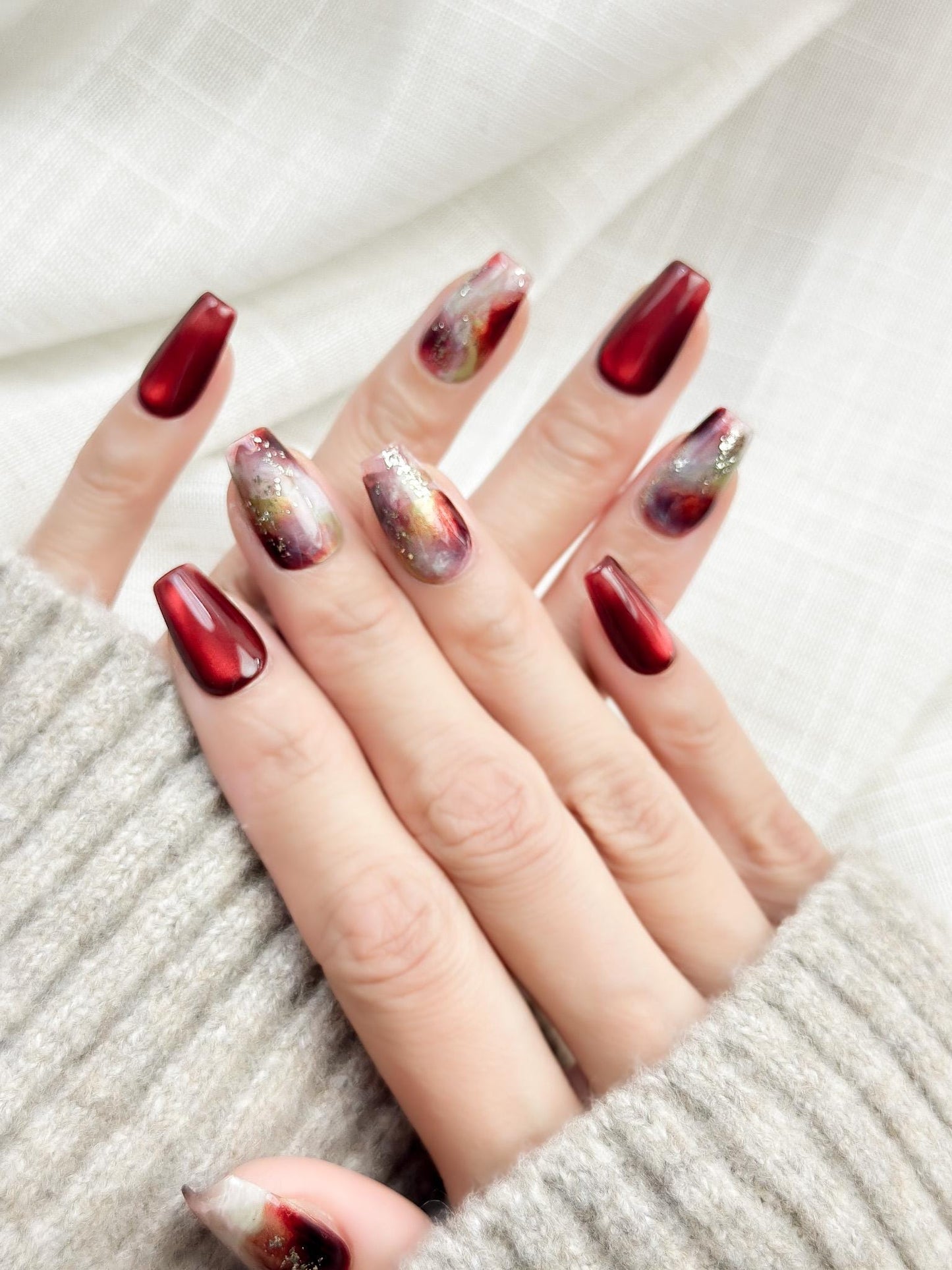 close up of hand with press-on nails in deep burgundy red with multi-color abstract art accents marble effect and silver glitter details, festive holiday nail styles