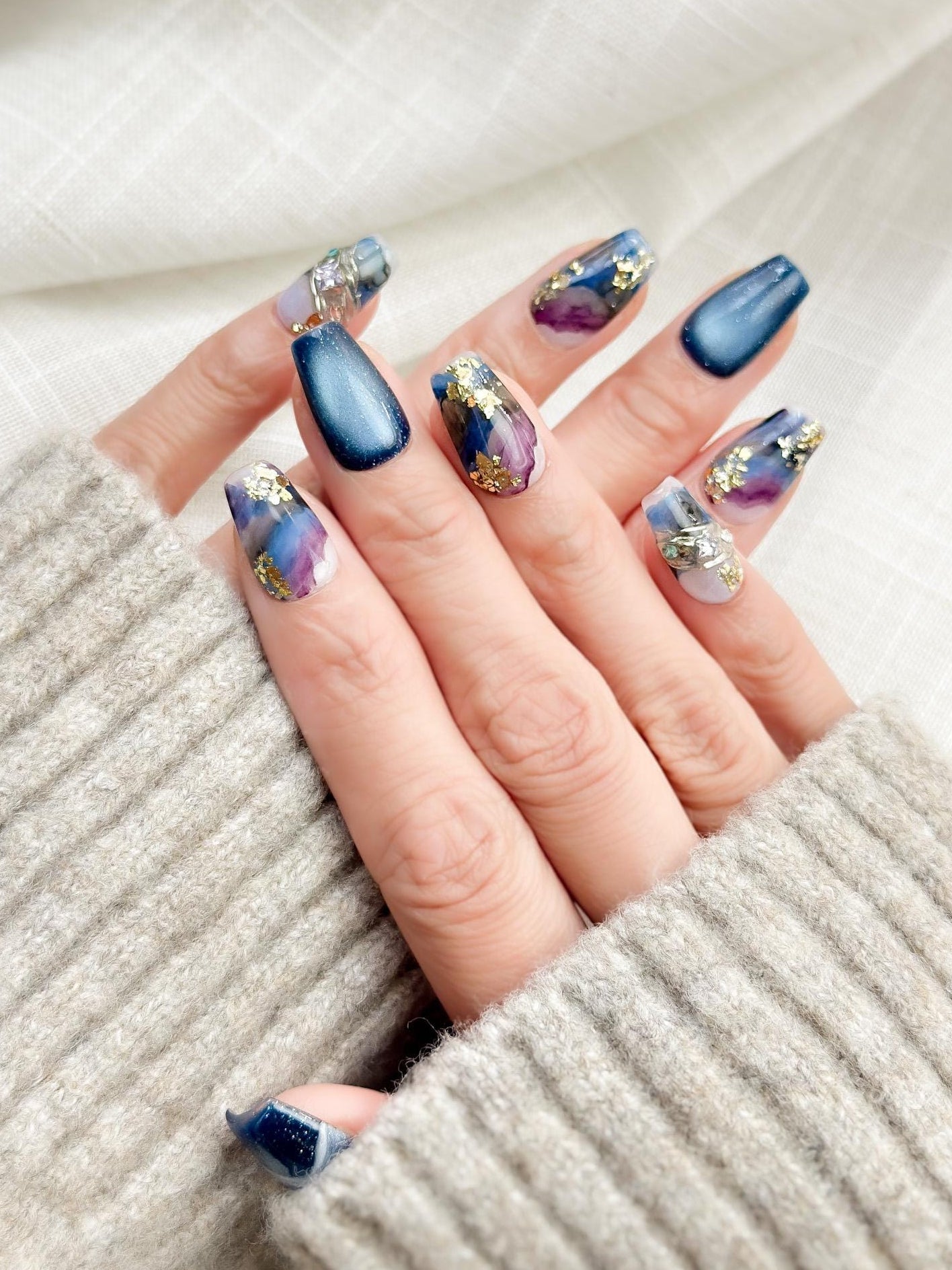 Hand with Blue galaxy press-on nails with starry cat-eye shimmer and watercolor marble accents in navy, lavender, and white, finished with gold foil details