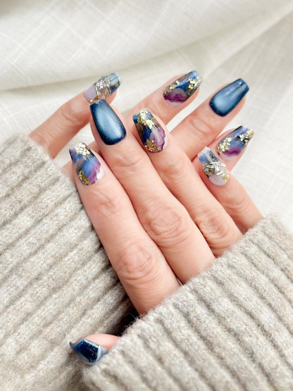 Hand with Blue galaxy press-on nails with starry cat-eye shimmer and watercolor marble accents in navy, lavender, and white, finished with gold foil details