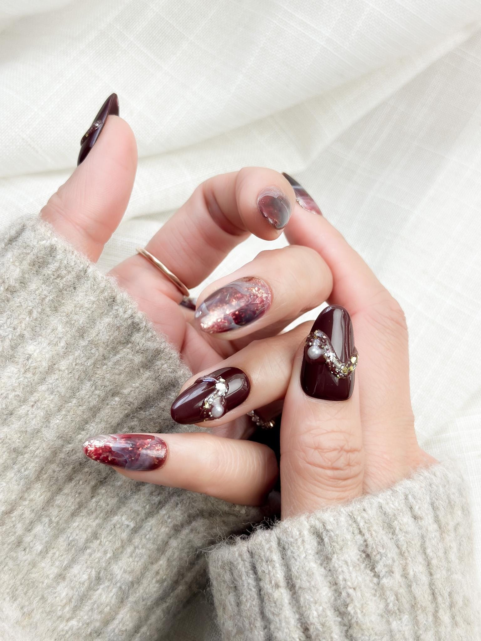 Hand with Almond-shaped burgundy and wine-red press-on nails with glossy finish, abstract marble art, gold glitter accents, and crystal pearl embellishments.