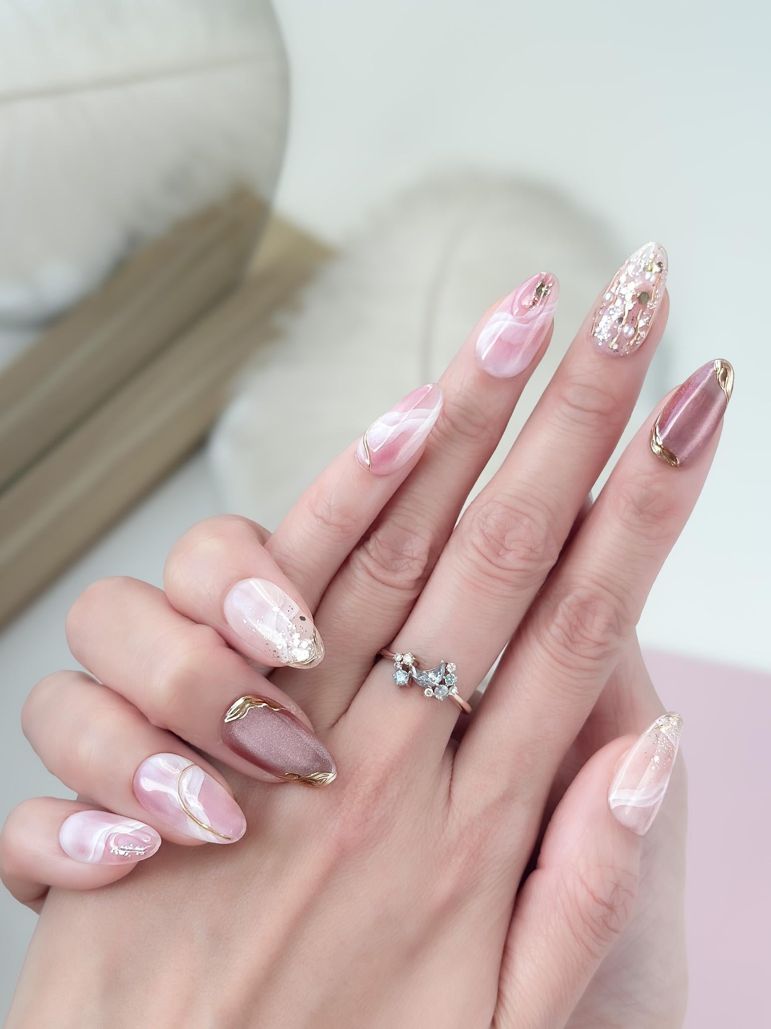 Close of of hand with Pink and nude luxury press-on nails with marble swirl designs, gold metallic accents, pearls, and glitter