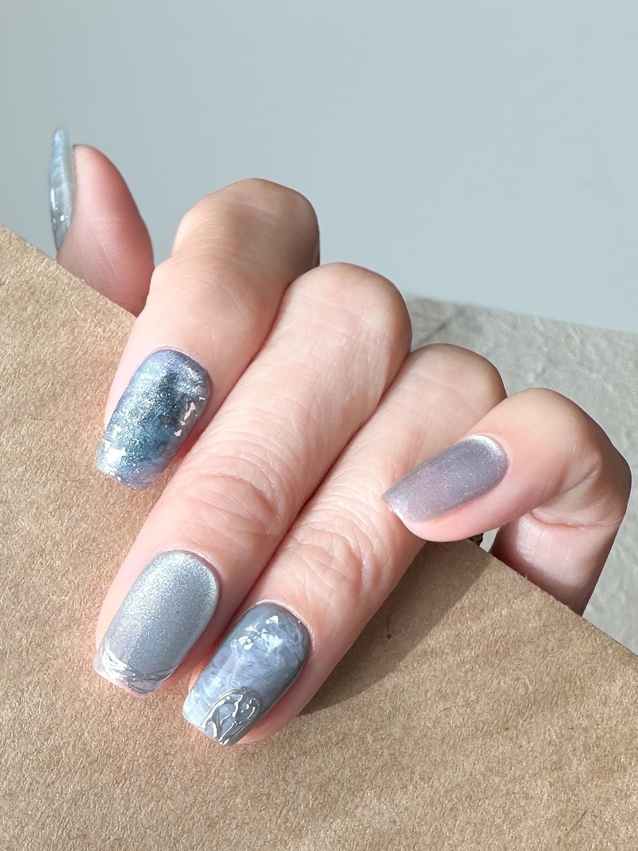 Hand with Blue and silver marble press-on nails with icy shimmer, metallic accents, and scattered silver foil flakes. Winter-inspired design featuring frosted blue hues and glossy snow-like details