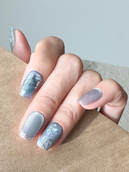Hand with Blue and silver marble press-on nails with icy shimmer, metallic accents, and scattered silver foil flakes. Winter-inspired design featuring frosted blue hues and glossy snow-like details