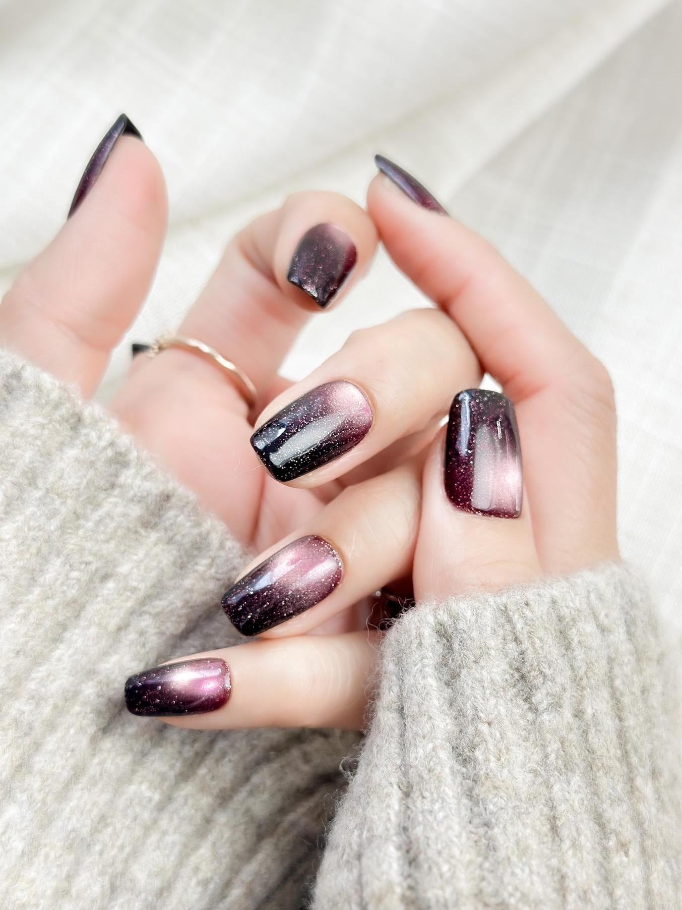 Hand with Short square press-on nails in a deep plum purple cat-eye shimmer with black gradient edges and silver sparkle accents