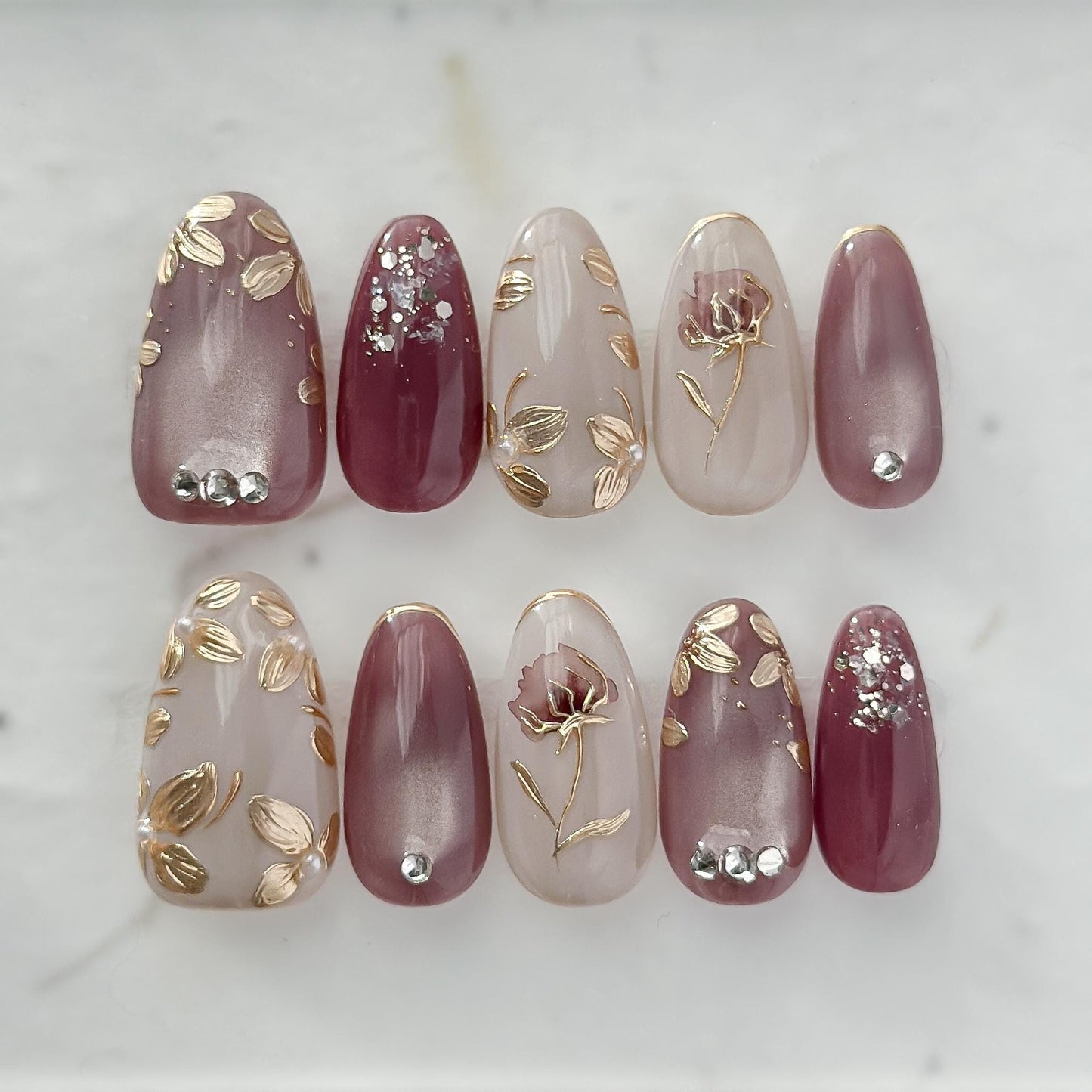 Almond shaped Press-on nails in deep plum and mauve tones with gold leaf accents, metallic floral designs, and silver gemstone details
