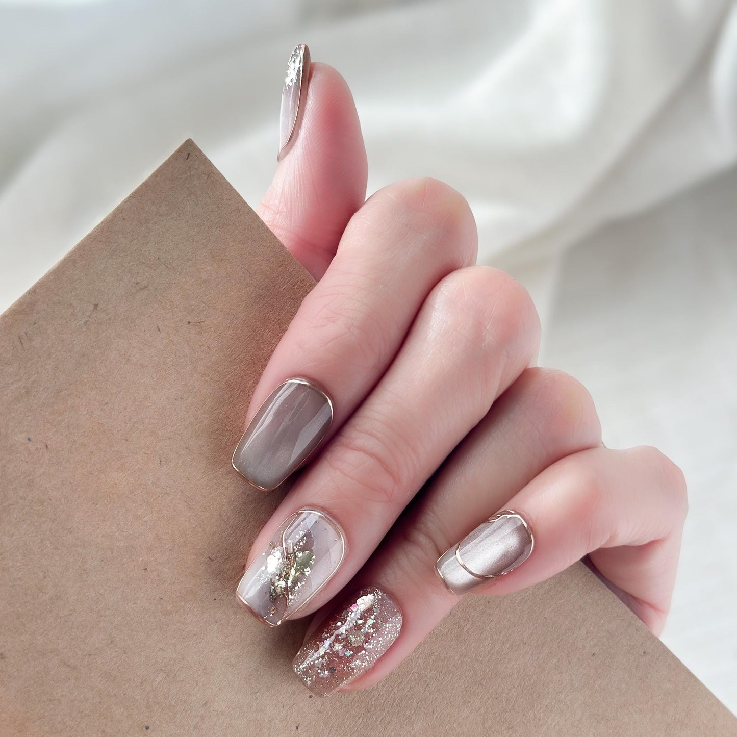 Close-up of hands with nude and taupe press-on nails with gold foil accents, rose gold glitter, and marble details