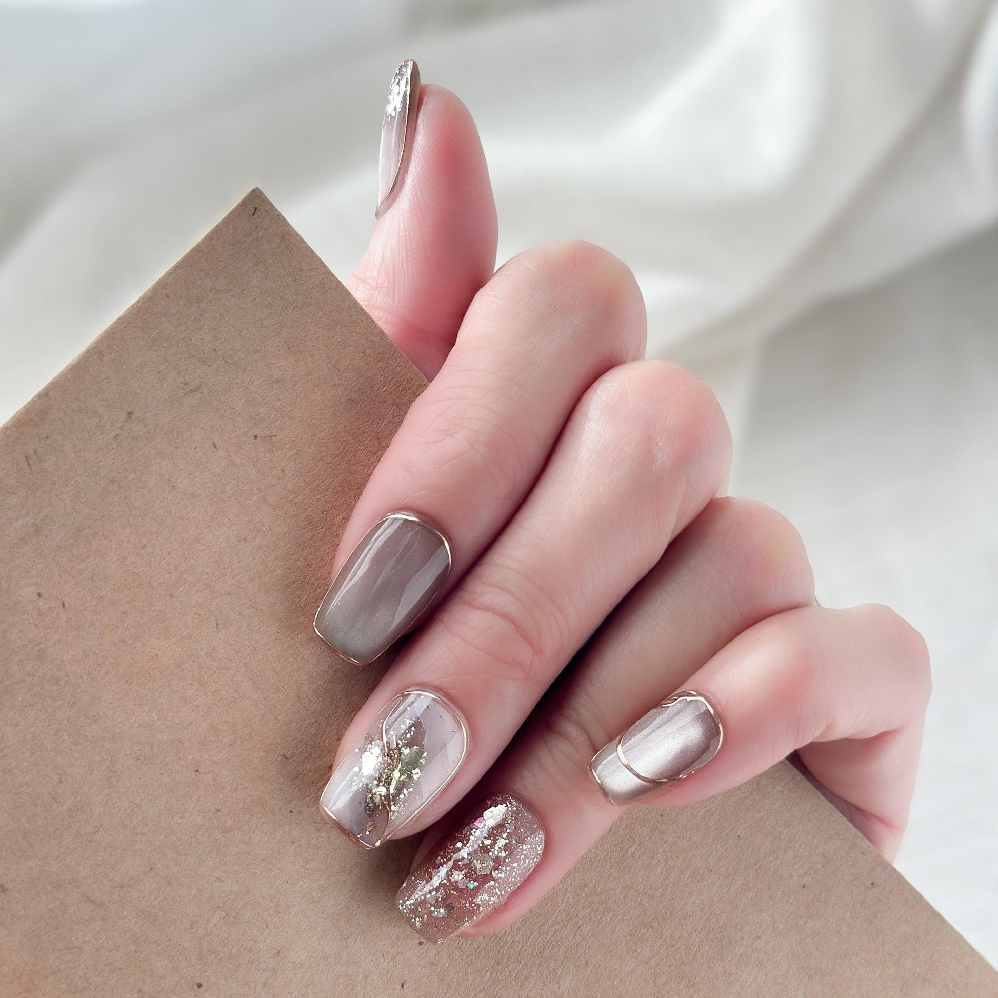 Close-up of hands with nude and taupe press-on nails with gold foil accents, rose gold glitter, and marble details