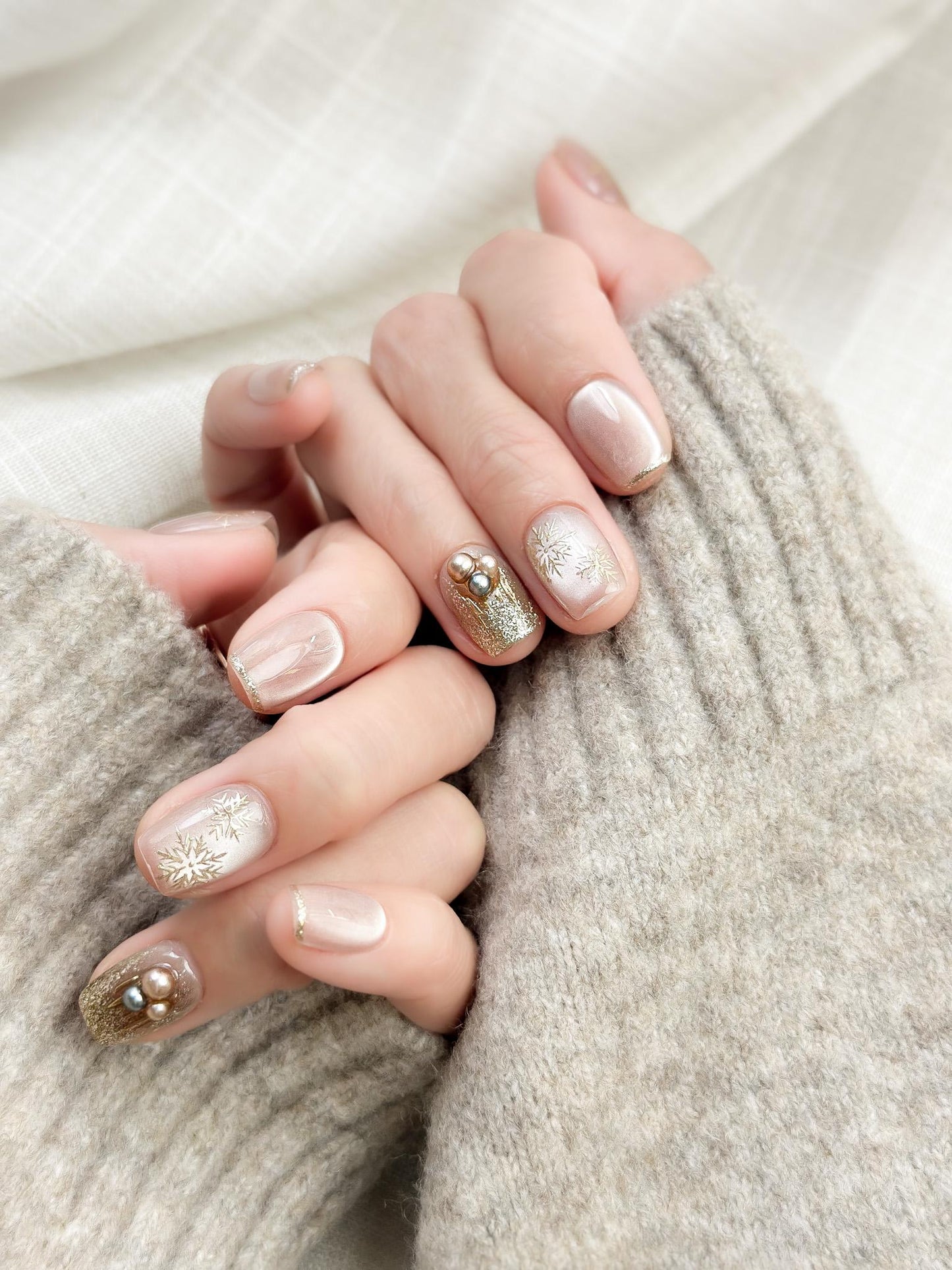 Hand with nude pink press-on nails with gold snowflake designs and glitter accents, featuring pearl embellishments for a festive holiday look