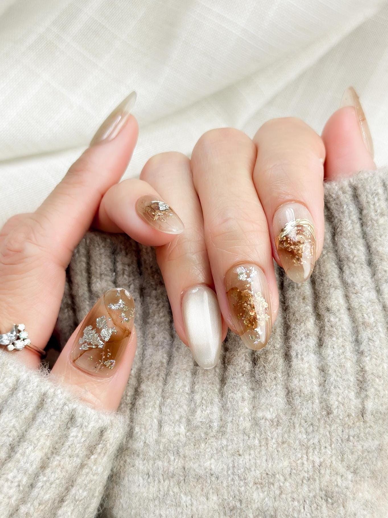 Close-up of almond-shaped nude and caramel brown press-on nails with gold foil accents and subtle shimmer, styled against a soft beige knit sweater