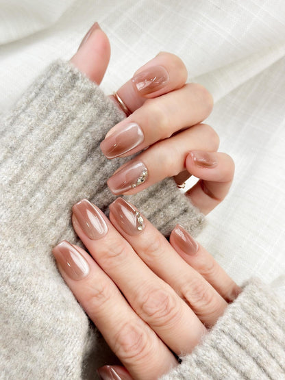 Close-up of a hand wearing glossy nude-brown press-on nails with a subtle cat-eye shimmer, featuring a gold leaf and crystal accent on one nail. The manicure is styled against a soft gray knit sweater for an elegant, cozy look
