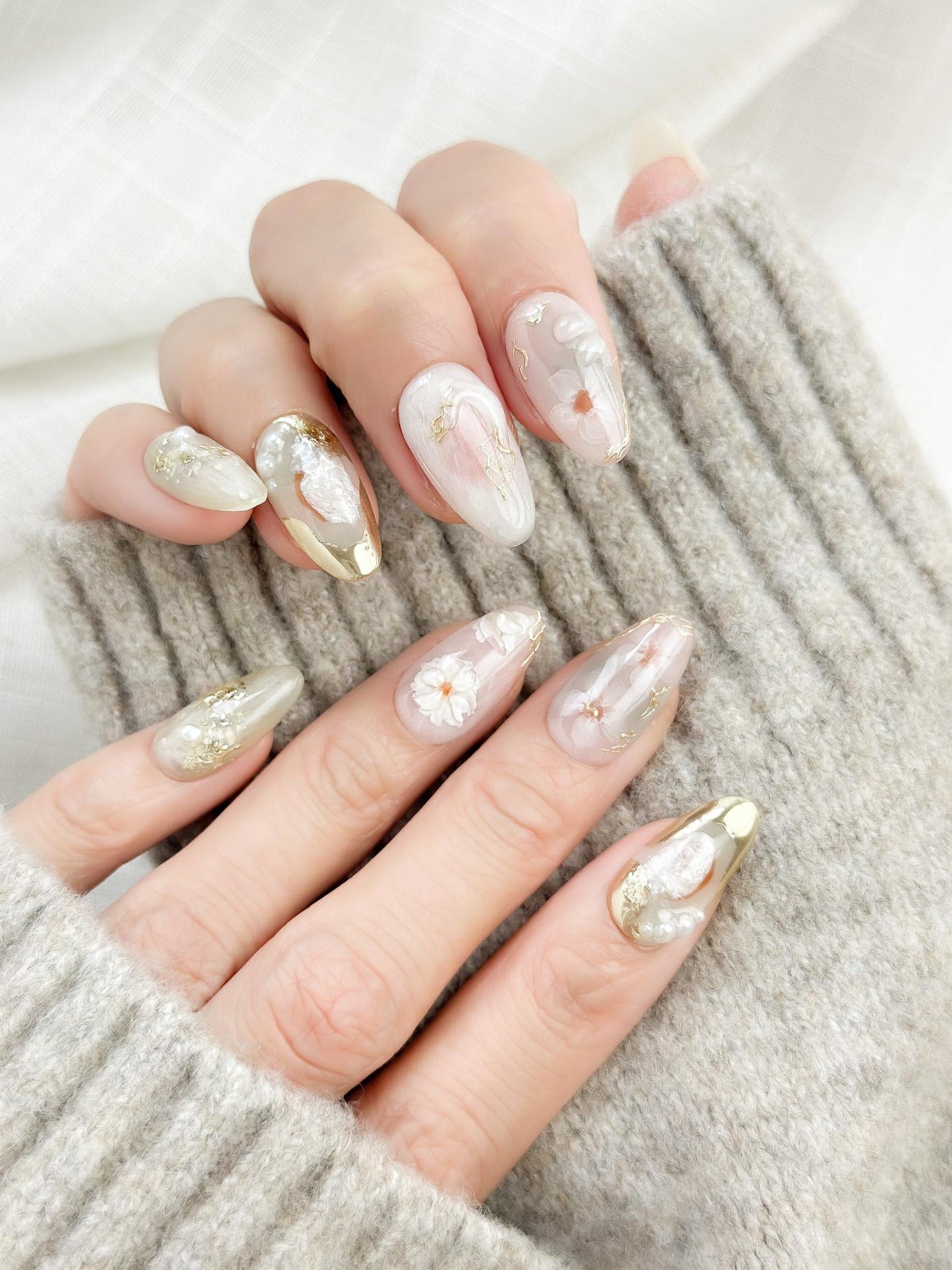 Almond-shaped white and pearlescent press-on nails with 3D floral accents, gold chrome details, pearl embellishments, and soft milky marble art