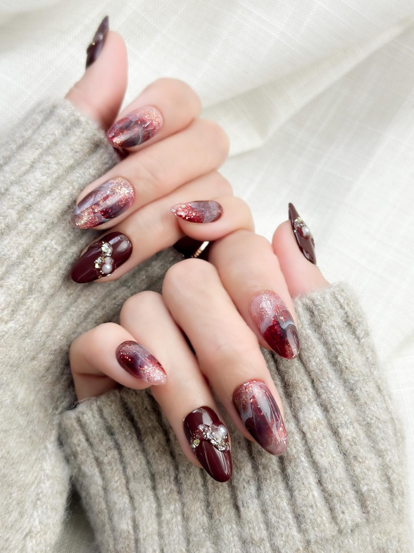 Hand with Almond-shaped burgundy and wine-red press-on nails with glossy finish, abstract marble art, gold glitter accents, and crystal pearl embellishments