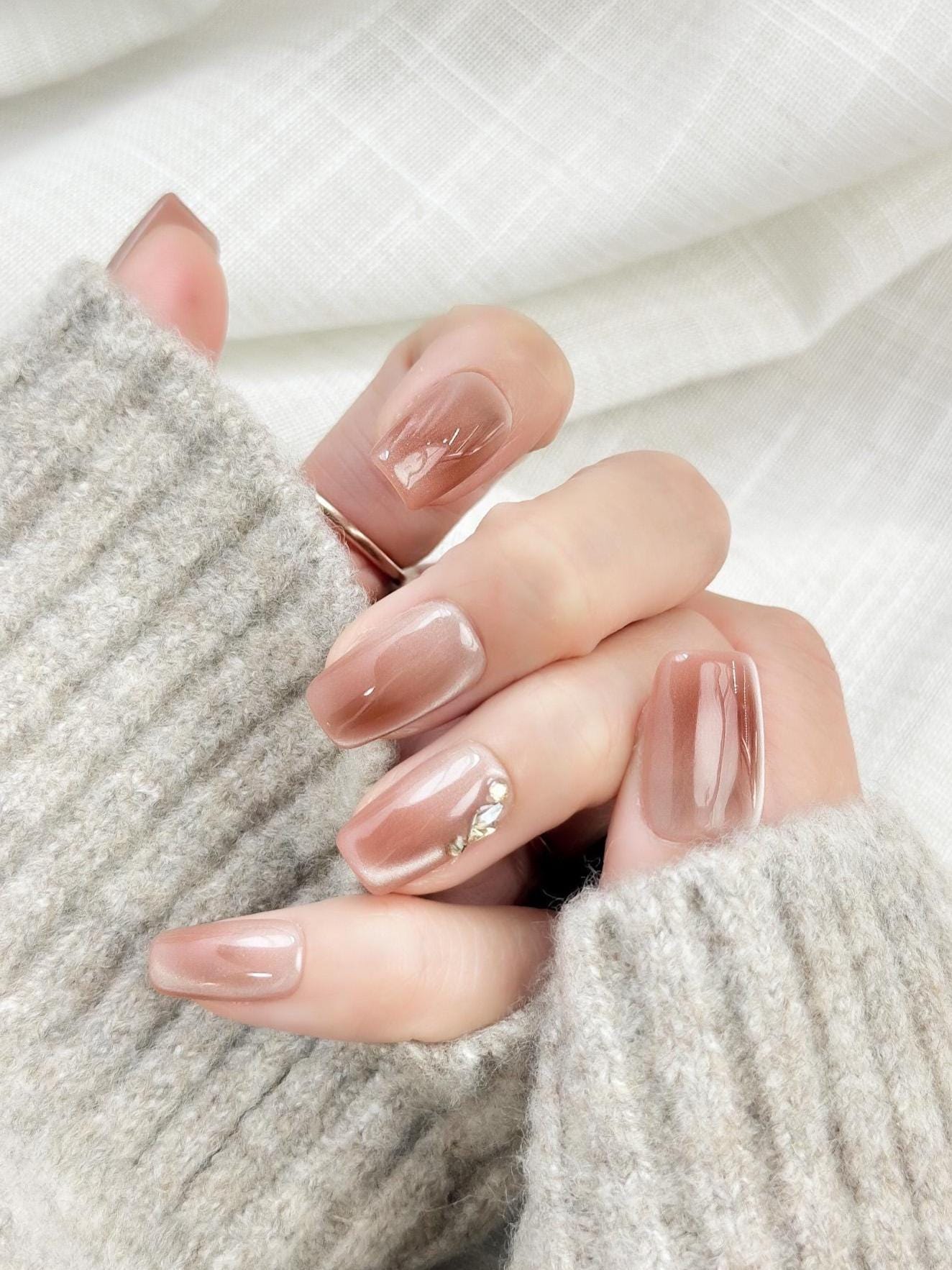 Close-up of a hand wearing glossy nude-brown press-on nails with a subtle cat-eye shimmer, featuring a gold leaf and crystal accent on one nail. The manicure is styled against a soft gray knit sweater for an elegant, cozy look