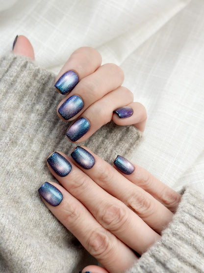 Hand with Short square shaped cat-eye press-on nails in shifting blue, purple, and teal galaxy shimmer