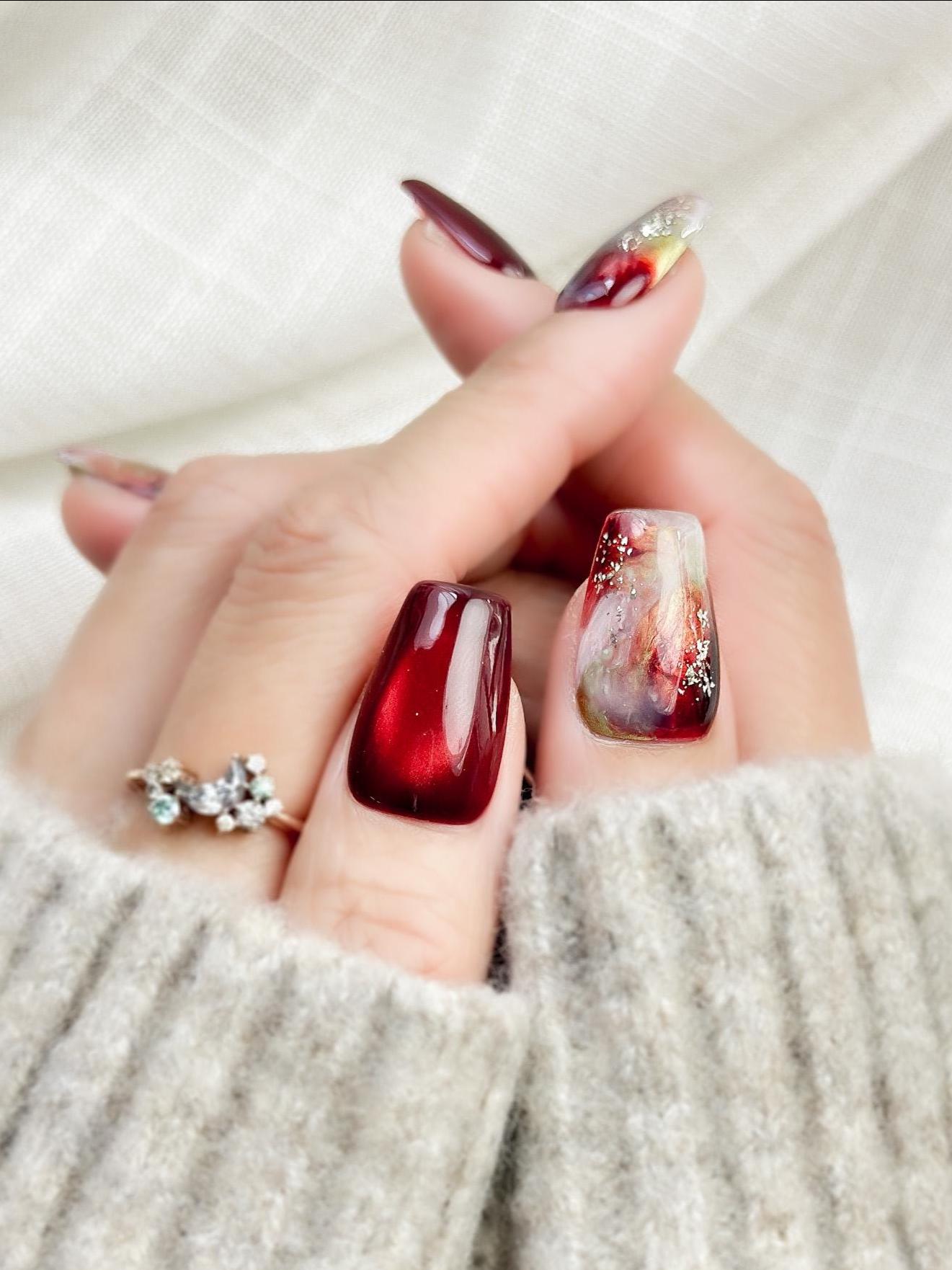 close up of hand with press-on nails in deep burgundy red with multi-color abstract art accents marble effect and silver glitter details, festive holiday style