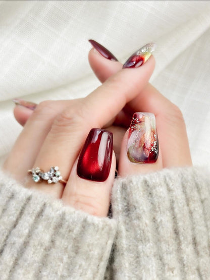 close up of hand with press-on nails in deep burgundy red with multi-color abstract art accents marble effect and silver glitter details, festive holiday style