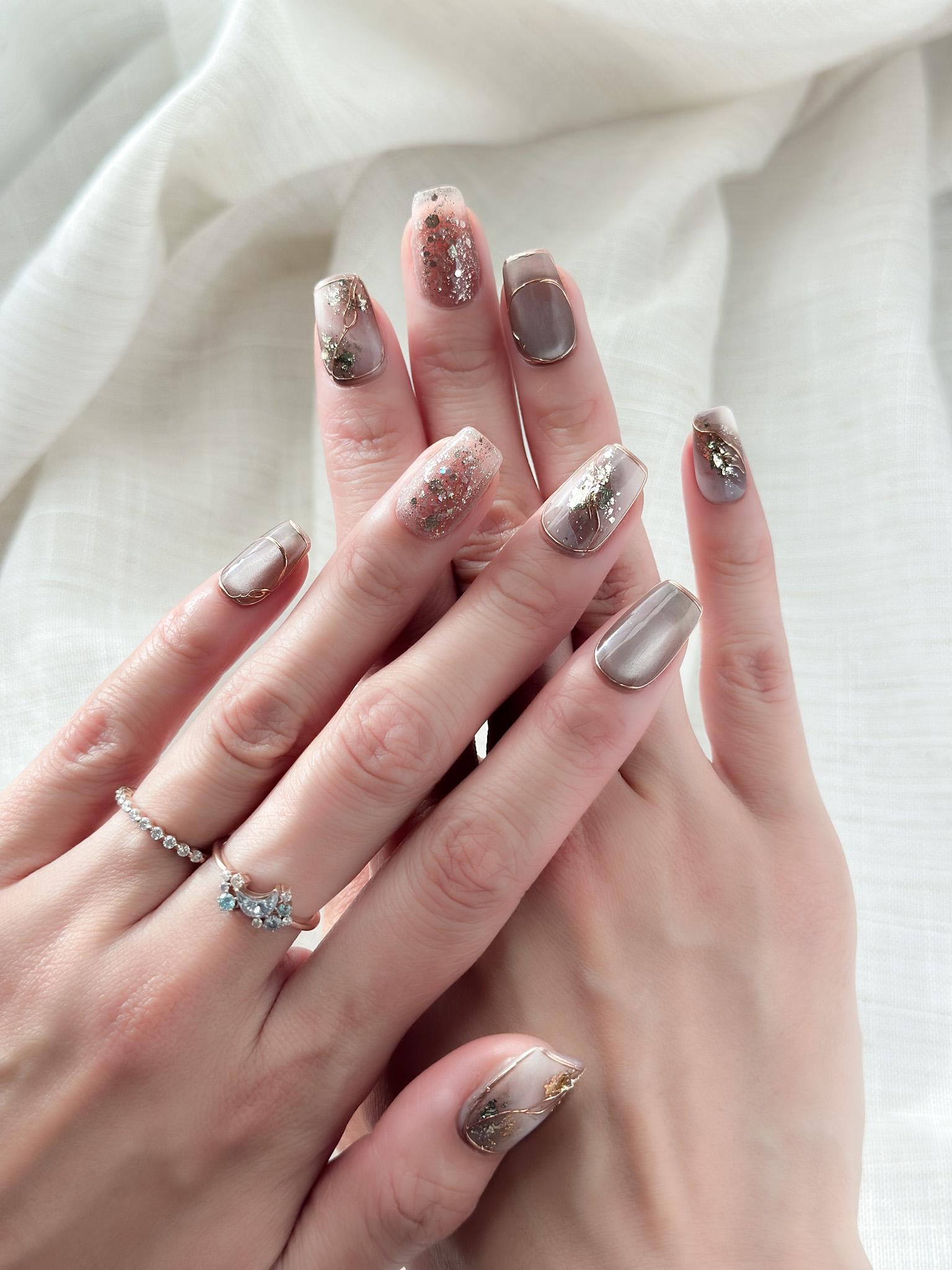 Close-up of hands with nude and taupe press-on nails with gold foil accents, rose gold glitter, and marble details