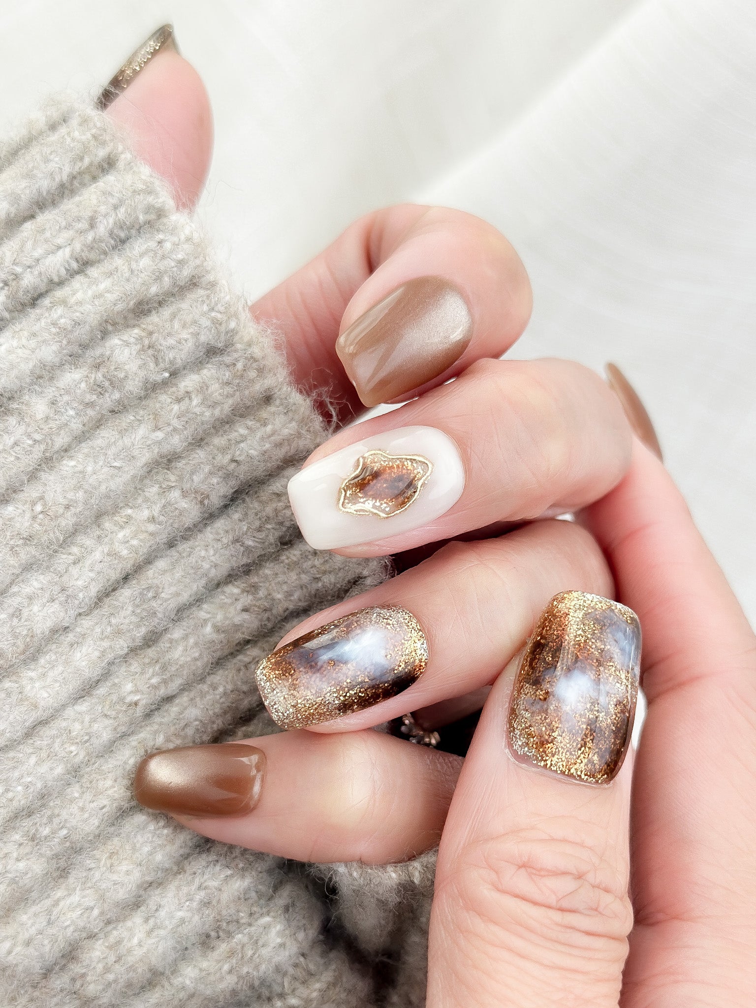 Hand with brown and ivory press-on nails in tortoise pattern, with gold foil detailing, and glossy cat-eye shimmer