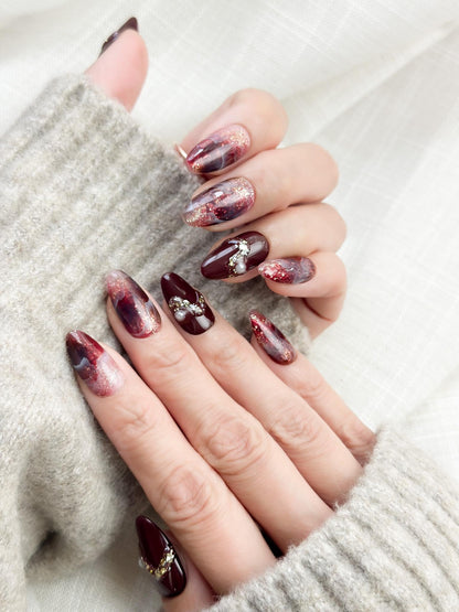 Hand with Almond-shaped burgundy and wine-red press-on nails with glossy finish, abstract marble art, gold glitter accents, and crystal pearl embellishments.