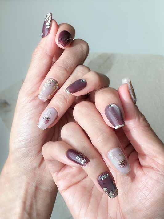 Hand with press-on nails in square coffin shape in deep plum and mauve tones with gold leaf accents, metallic floral designs, and silver gemstone details.