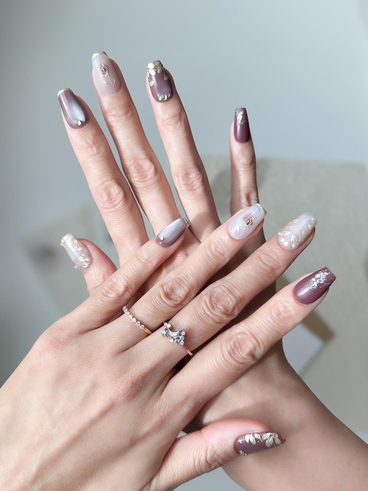Hand with press-on nails in square coffin shape in deep plum and mauve tones with gold leaf accents, metallic floral designs, and silver gemstone details