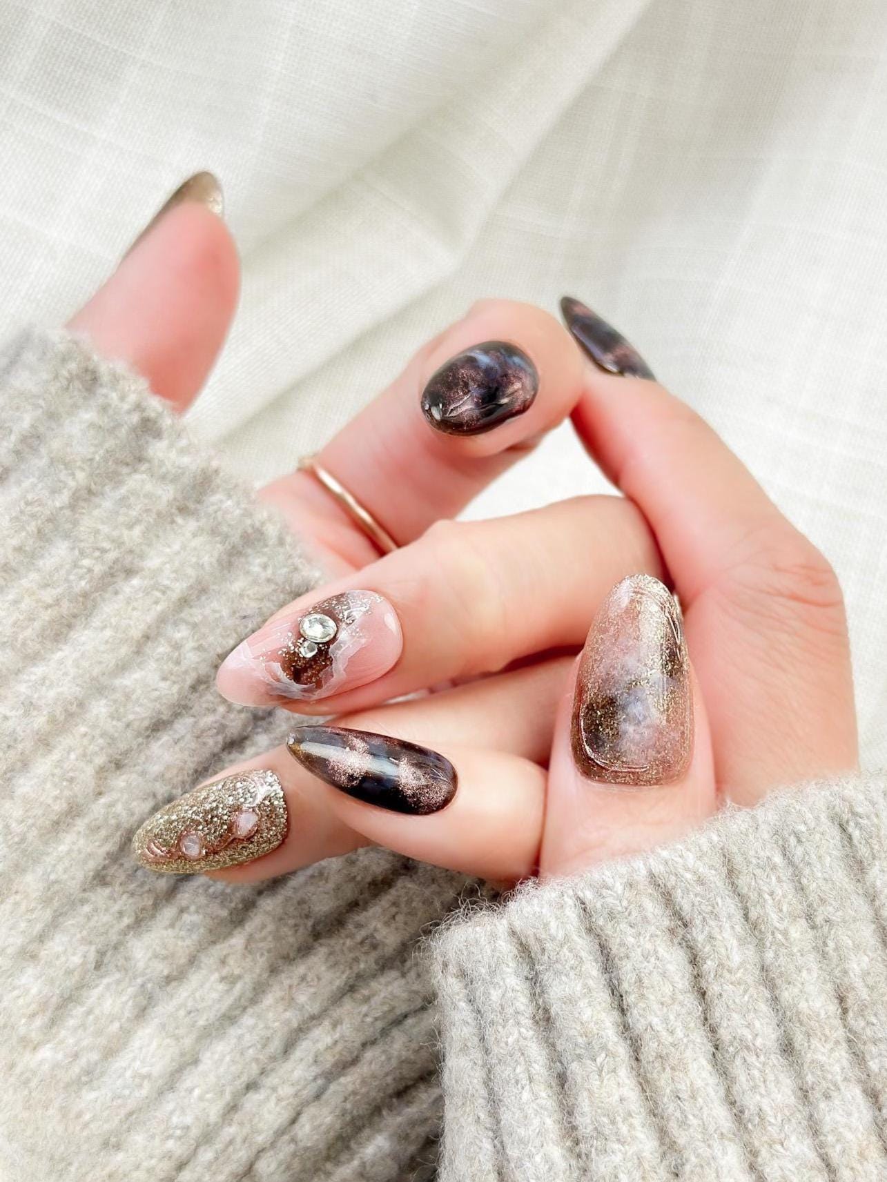 Hand with Almond-shaped luxury press-on nails in metallic brown and black cat-eye shimmer with pink marble accents, gold glitter, and pearl gemstone details