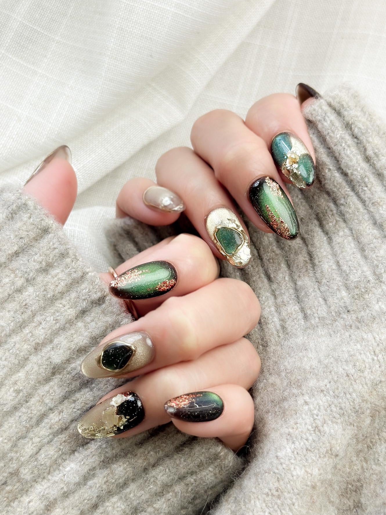 Hand with Green and gold gemstone-inspired press-on nails with cat-eye shimmer, glitter accents, gold foil detailing, and 3D crystal embellishments