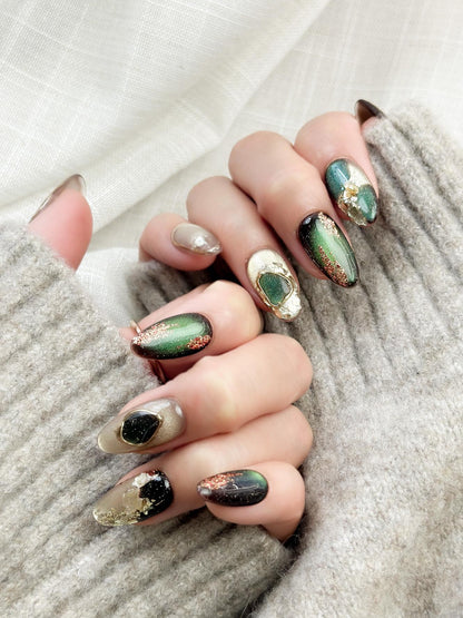 Hand with Green and gold gemstone-inspired press-on nails with cat-eye shimmer, glitter accents, gold foil detailing, and 3D crystal embellishments