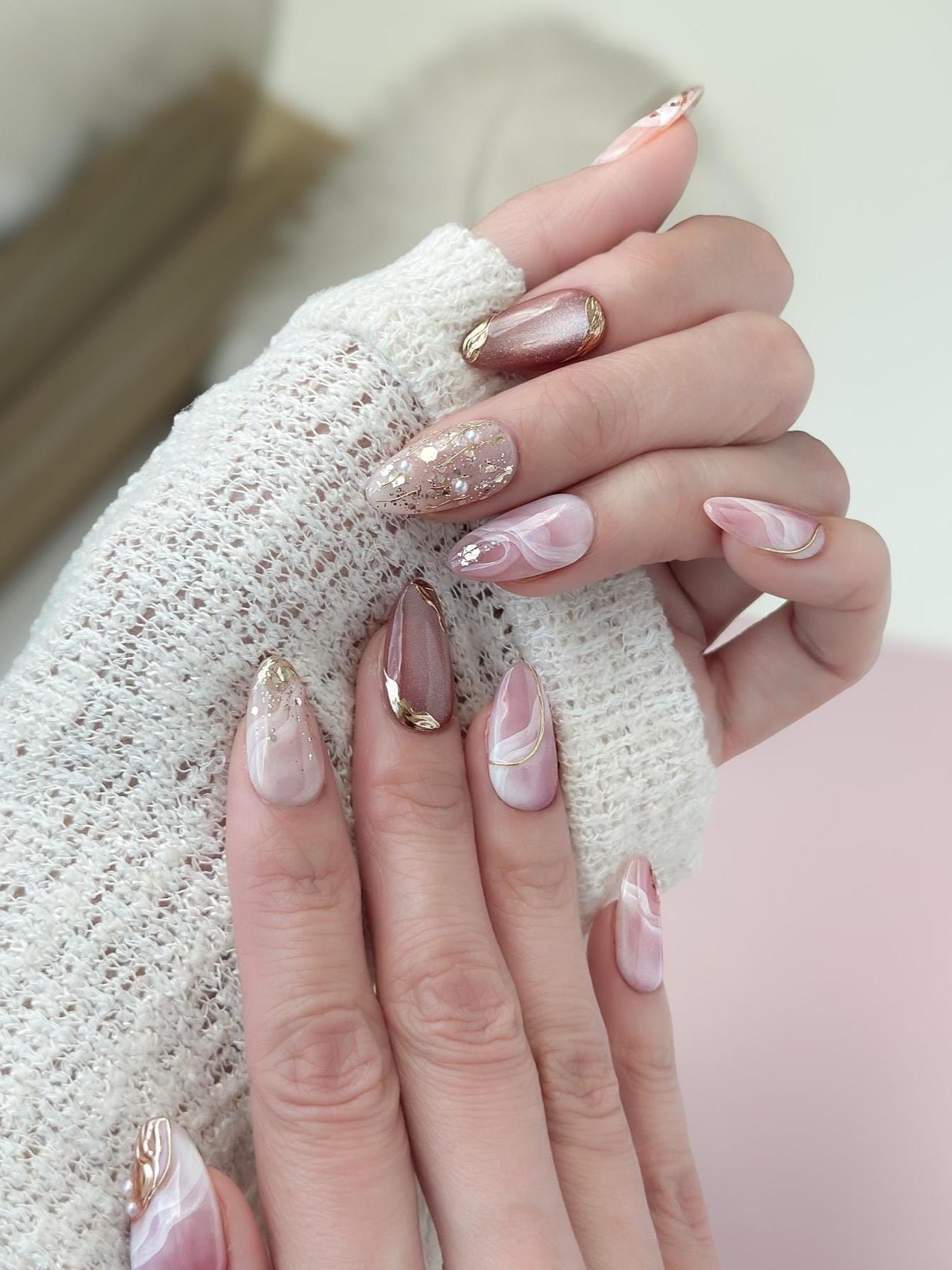 Close of of hand with Pink and nude luxury press-on nails with marble swirl designs, gold metallic accents, pearls, and glitter