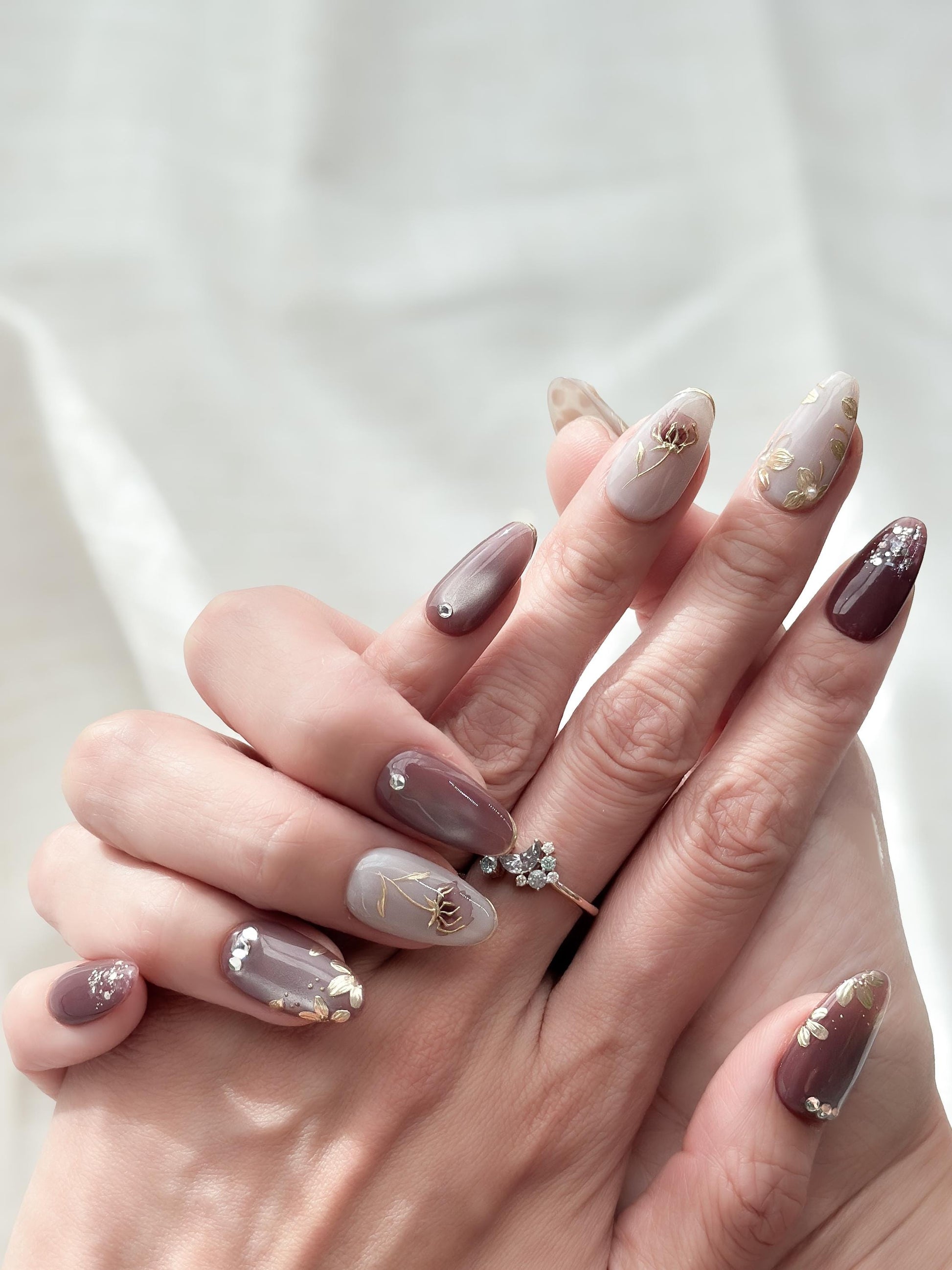 Hand with almond shaped Press-on nails in deep plum and mauve tones with gold leaf accents, metallic floral designs, and silver gemstone details