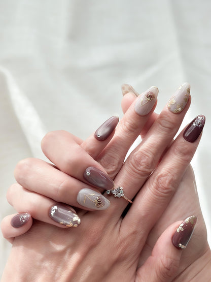 Hand with almond shaped Press-on nails in deep plum and mauve tones with gold leaf accents, metallic floral designs, and silver gemstone details