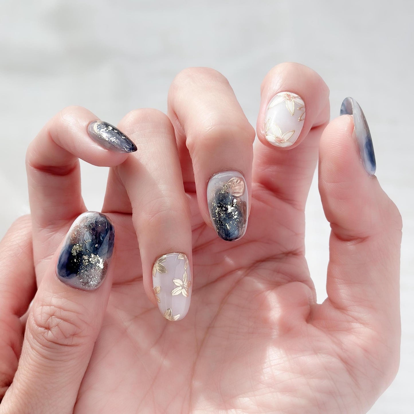 Hand with Oval-shaped press-on nails with deep navy and gold marble art, gold foil accents, and soft floral details.