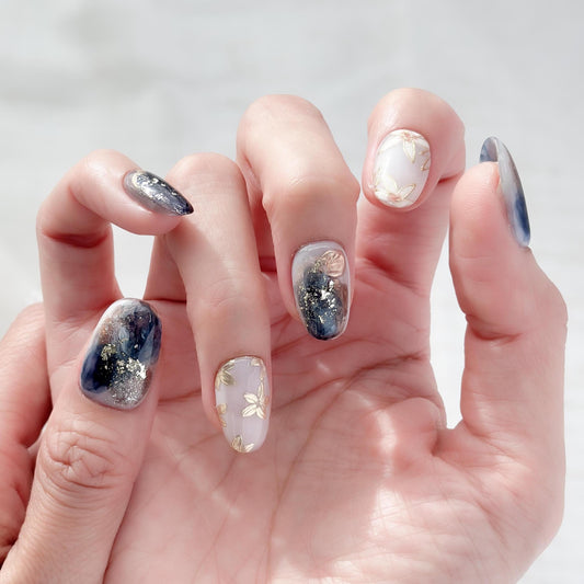 Hand with Oval-shaped press-on nails with deep navy and gold marble art, gold foil accents, and soft floral details.