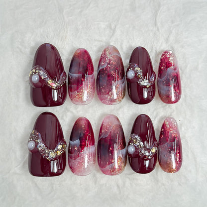 Almond-shaped burgundy and wine-red press-on nails with glossy finish, abstract marble art, gold glitter accents, and crystal pearl embellishments