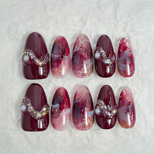 Almond-shaped burgundy and wine-red press-on nails with glossy finish, abstract marble art, gold glitter accents, and crystal pearl embellishments