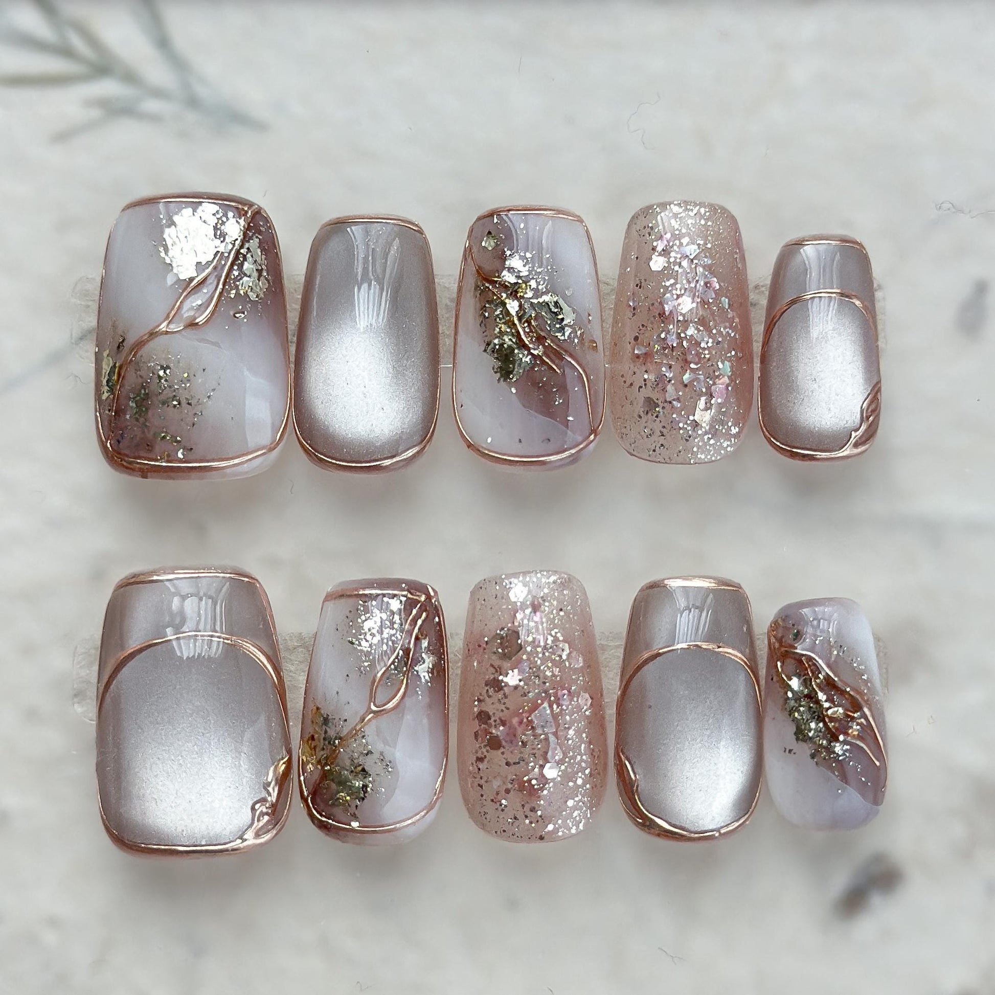 A set of nude and taupe press-on nails with gold foil accents, rose gold glitter, and marble details