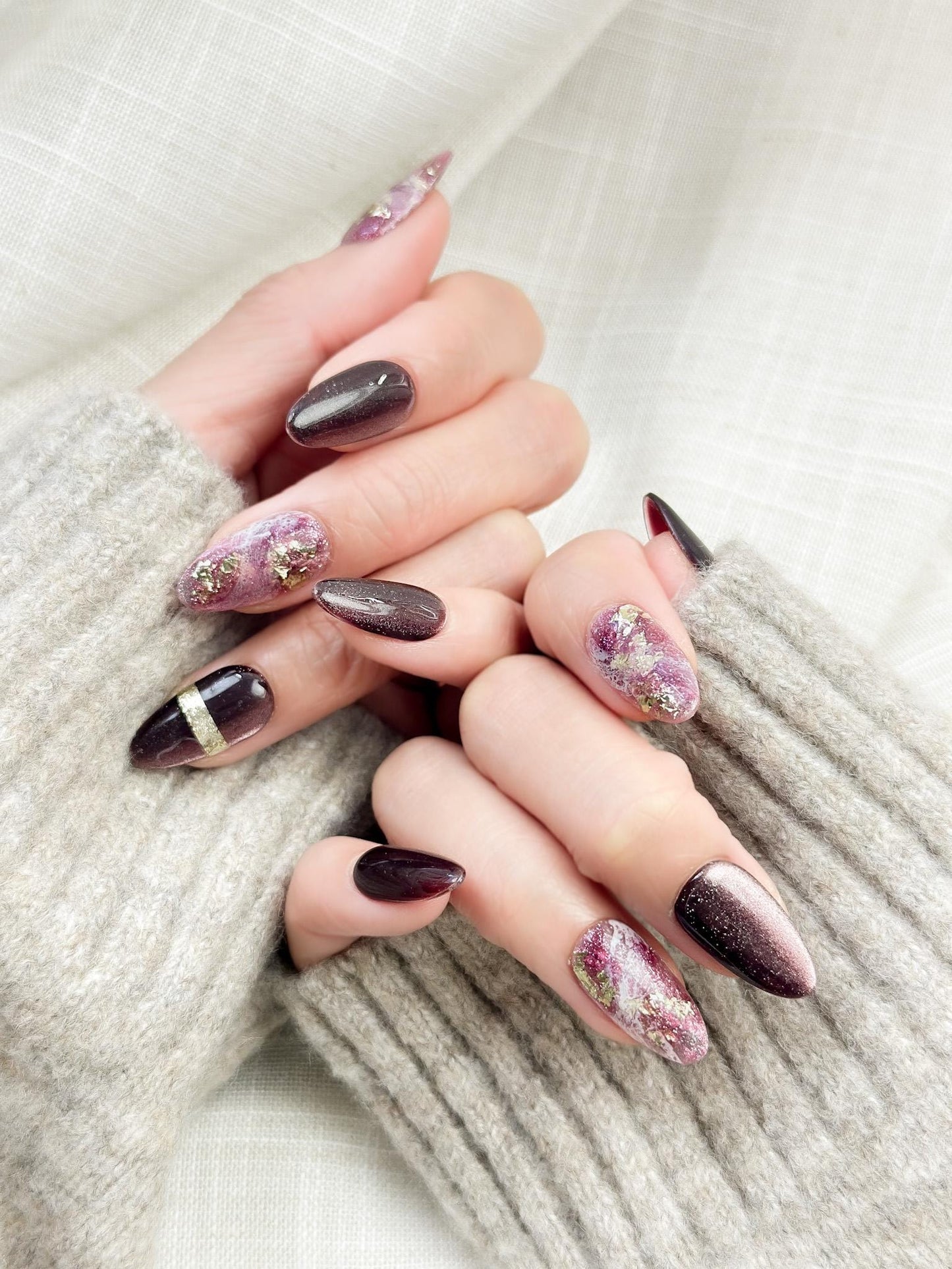 Hand with Almond-shaped press-on nails in deep plum and black cat-eye shimmer with pink marble accents, gold foil detailing, and glossy finish