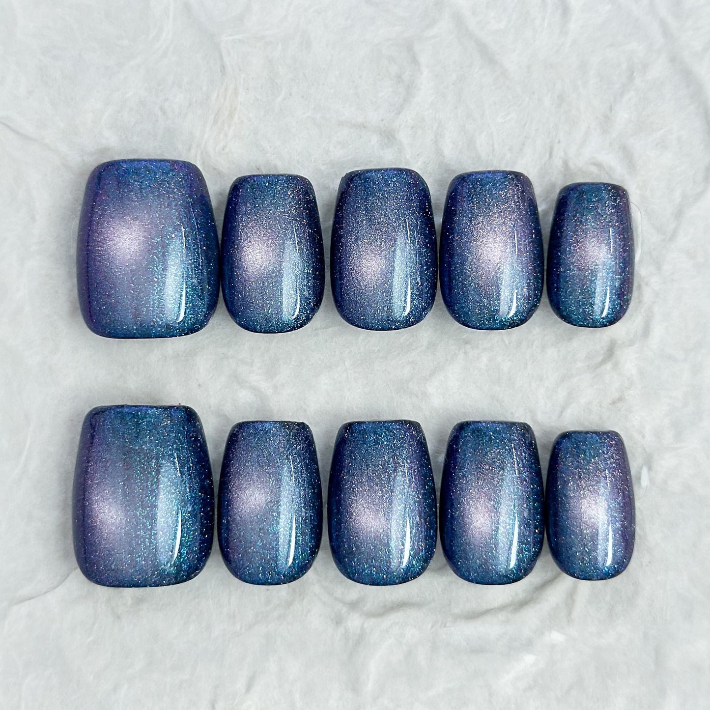 Short square shaped cat-eye press-on nails in shifting blue, purple, and teal galaxy shimmer