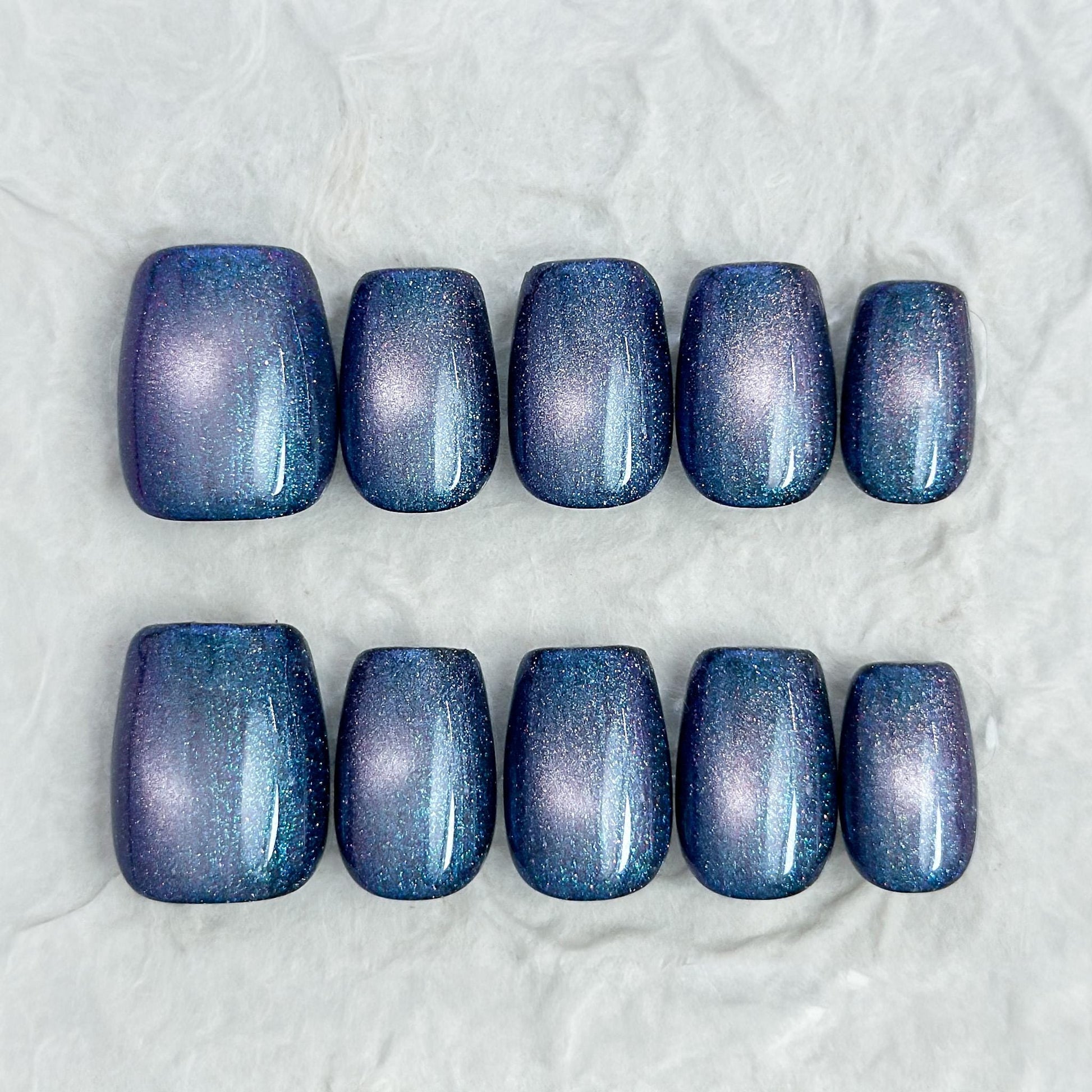 Short square shaped cat-eye press-on nails in shifting blue, purple, and teal galaxy shimmer