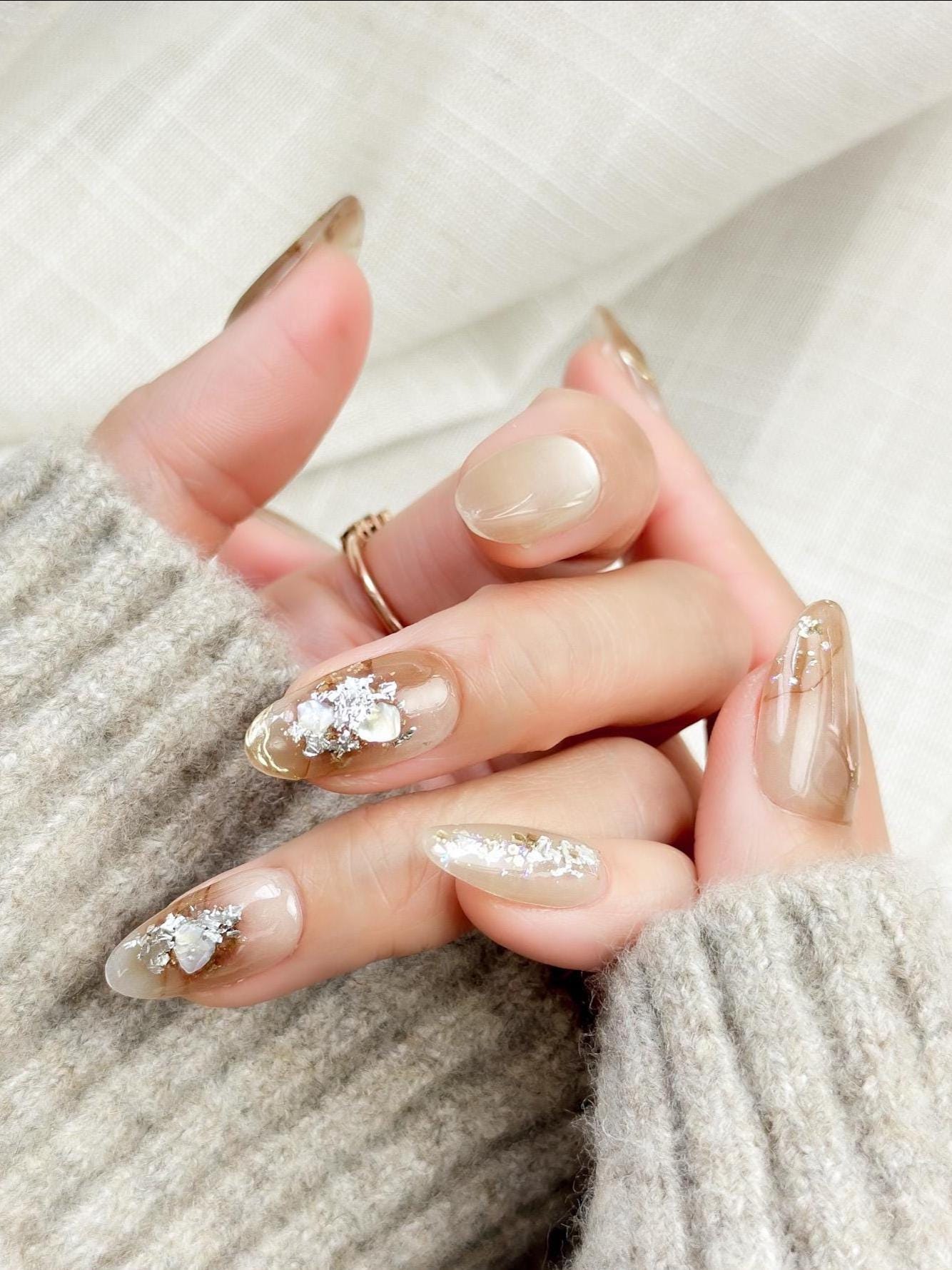Close-up of almond-shaped nude and caramel brown press-on nails with gold foil accents and subtle shimmer, styled against a soft beige knit sweater.